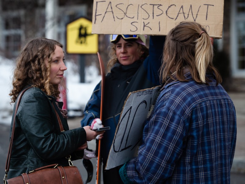 Three people stand outside in winter clothes, holding protest signs. One sign partially reads "Fascists Can't Ski." Snow is visible in the background.