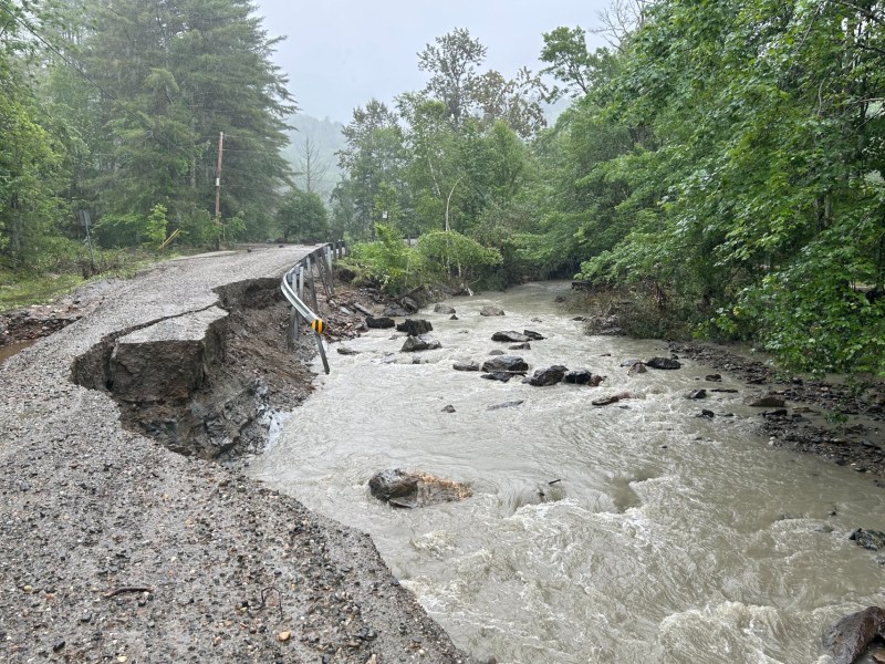 A washed-out road next to a swollen stream after heavy rainfall, with sections of the pavement collapsed into the water. Dense trees surround the area.