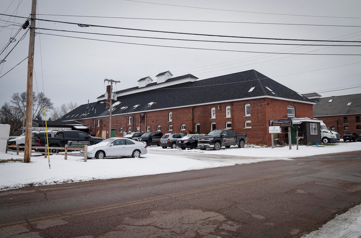 Large brick building with a dark roof, surrounded by parked cars. Snow covers the ground and rooftops. Power lines are visible overhead. A sign near the building indicates a business entrance.