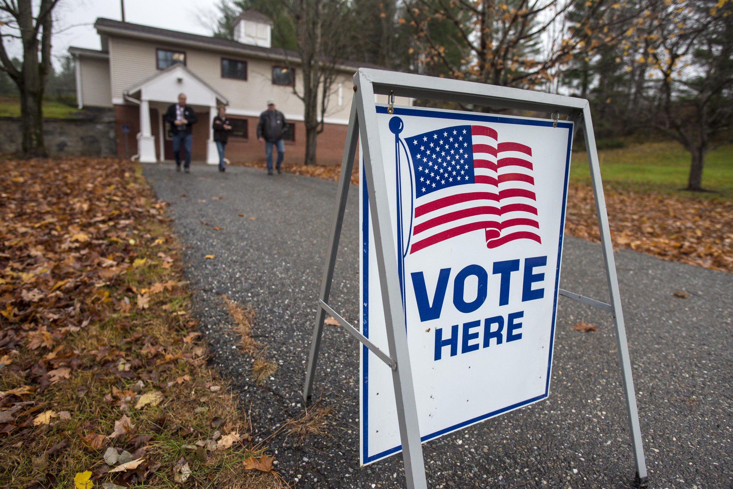 A "Vote Here" sign with an American flag is posted on a path leading to a building. Three people walk away from the building. Fallen leaves are scattered on the ground.