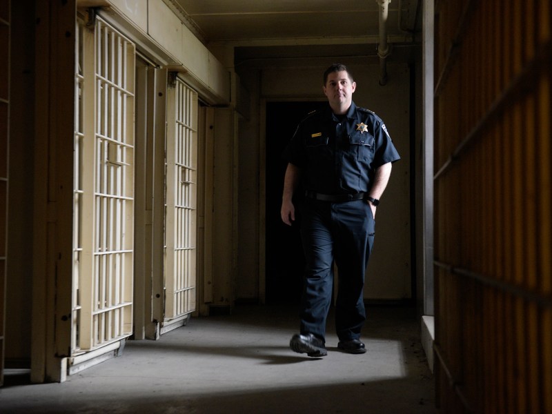 A correctional officer walks through a dimly lit hallway lined with jail cells.
