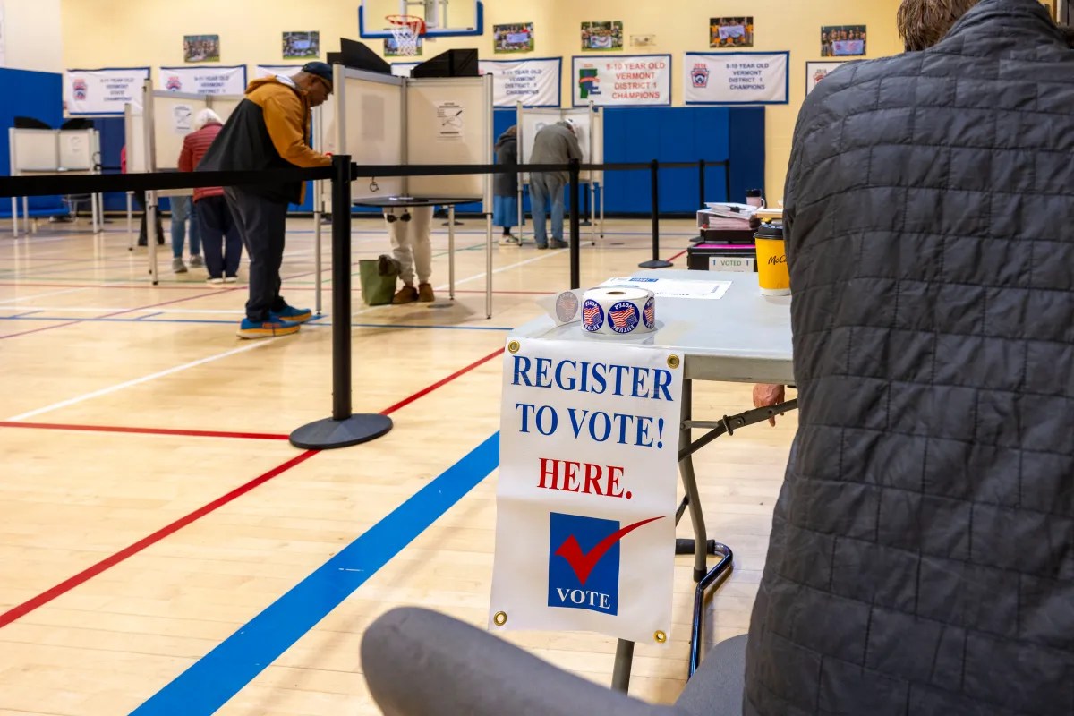 People voting in a gymnasium, with a "Register to Vote Here" sign on a table in the foreground.