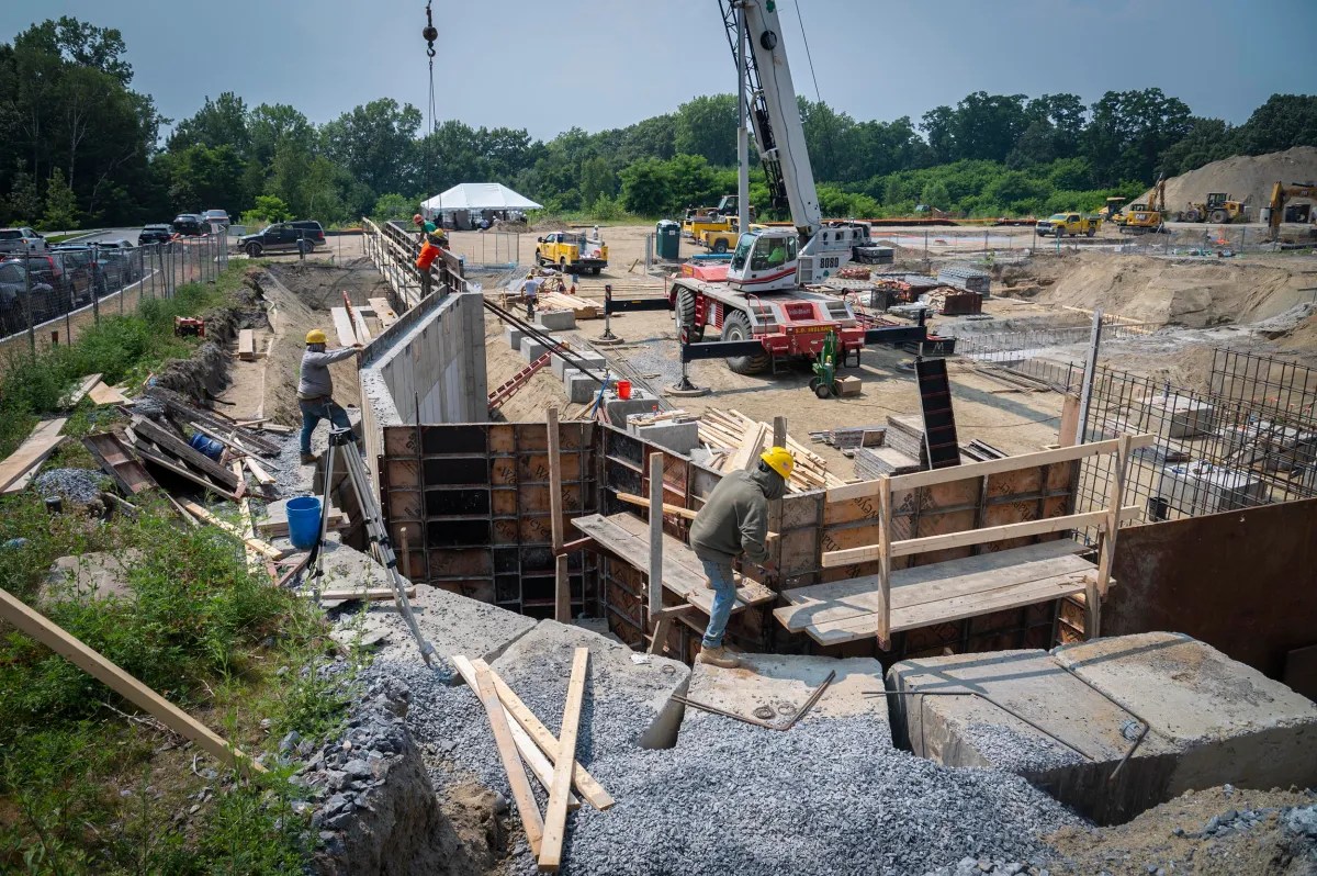 Construction workers wearing safety gear work on a building foundation at an active construction site with cranes, machinery, and materials visible.