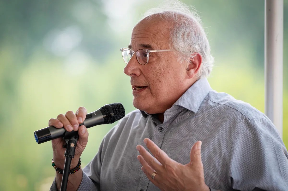 An older man with gray hair, glasses, and a light blue shirt speaks into a microphone while gesturing with his hand outdoors.