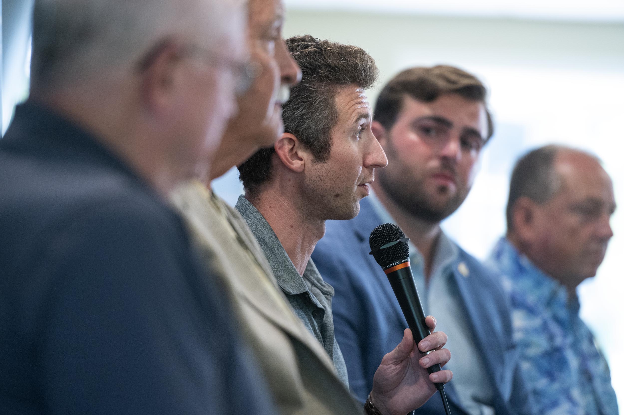 A man speaks into a microphone while seated in a row with four other men during a panel discussion.