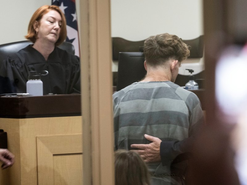 A judge sits at her bench while a person in a striped prison uniform stands before her, accompanied by another individual.