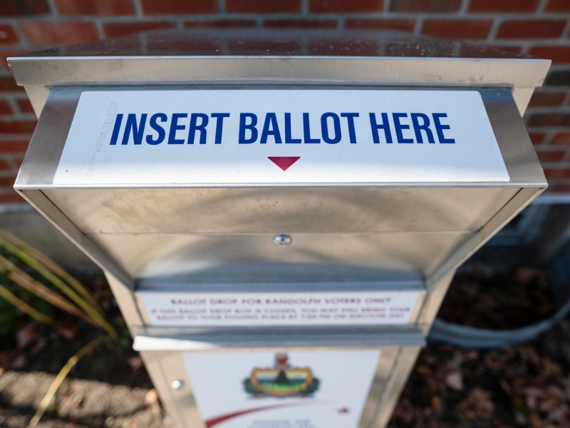 A ballot drop box with a sign reading "Insert Ballot Here" is positioned outside against a brick wall.