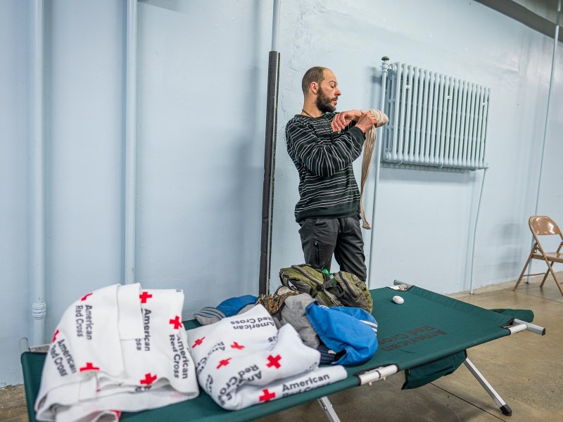Man standing near a cot with Red Cross blankets in a shelter, putting on a jacket.