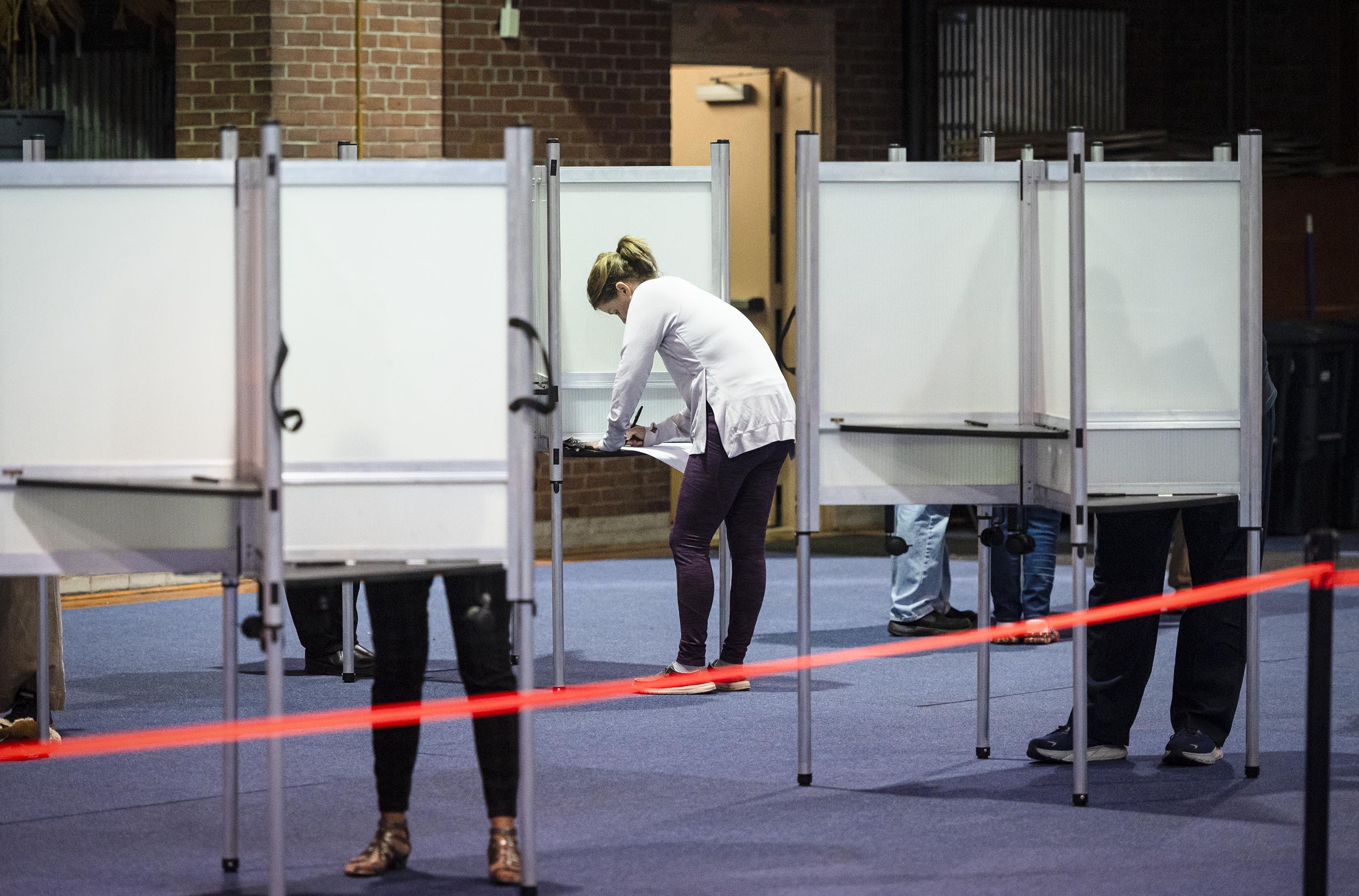 People standing at voting booths in a voting station with a red tape barrier in the foreground.