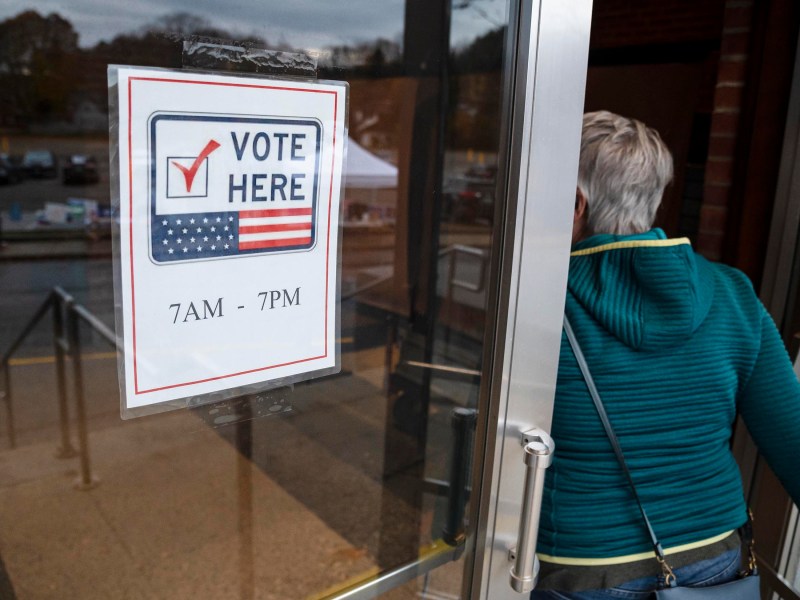 Person entering a polling station with a "Vote Here" sign on the door, indicating voting hours from 7 AM to 7 PM.