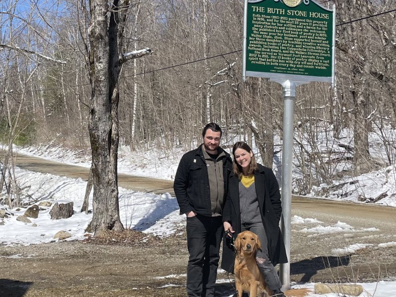 Two people stand with a dog on a snowy roadside next to a historical marker for "The Ruth Stone House." Bare trees and a dirt road are in the background.