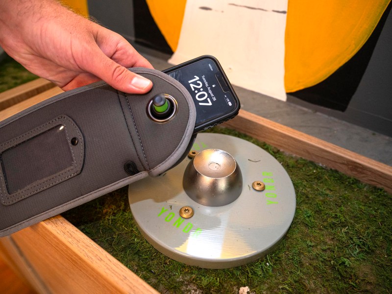 A person scans a round metal object labeled "YOND" with a smartphone inside a grey pouch, placed on a grassy surface in a wooden enclosure.