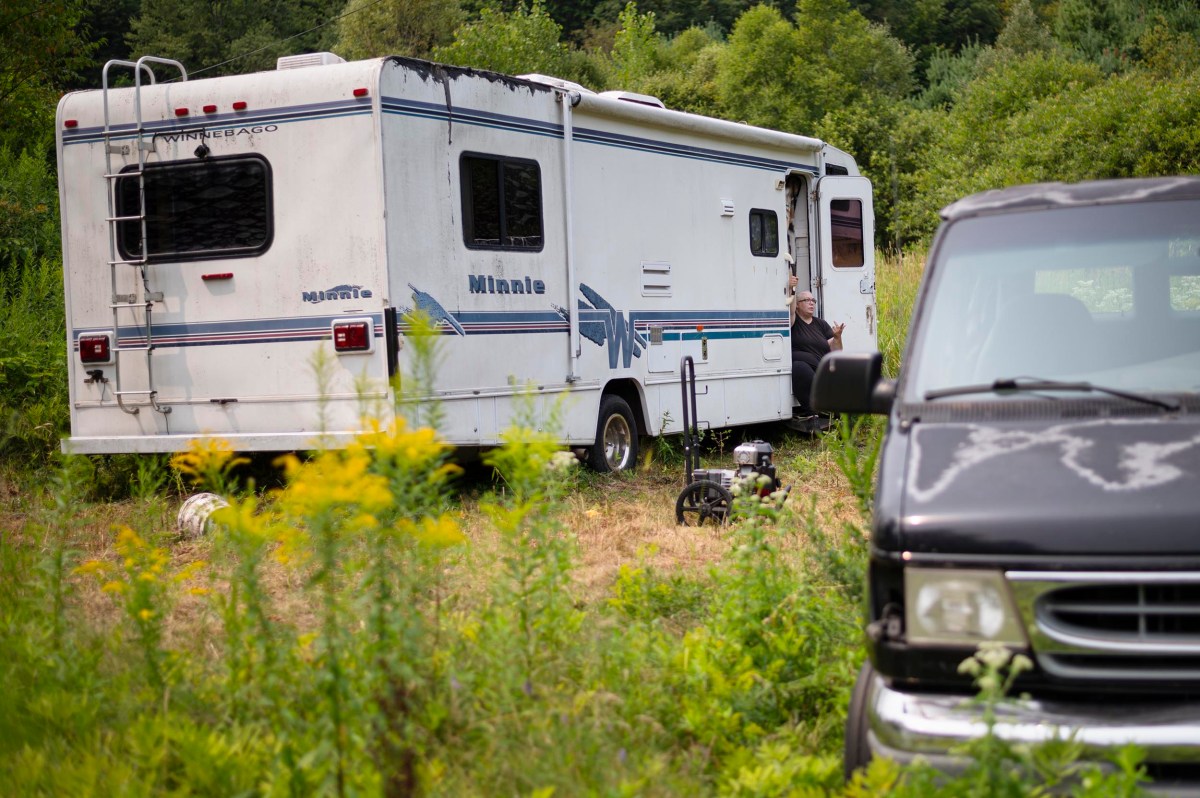 A white Winnebago RV is parked on grass near dense foliage, with a black van in the foreground and a person standing at the RV’s open door.