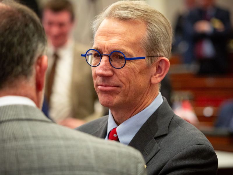 A man in a suit with blue-rimmed glasses is engaged in conversation with another person at an indoor event.