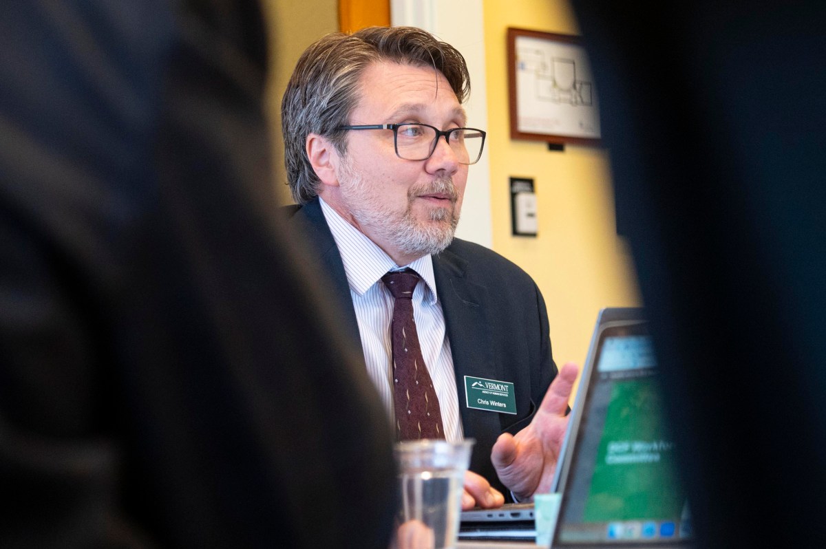 A man wearing glasses and a suit speaks during a meeting, seated at a table with laptops and a glass of water in front of him.