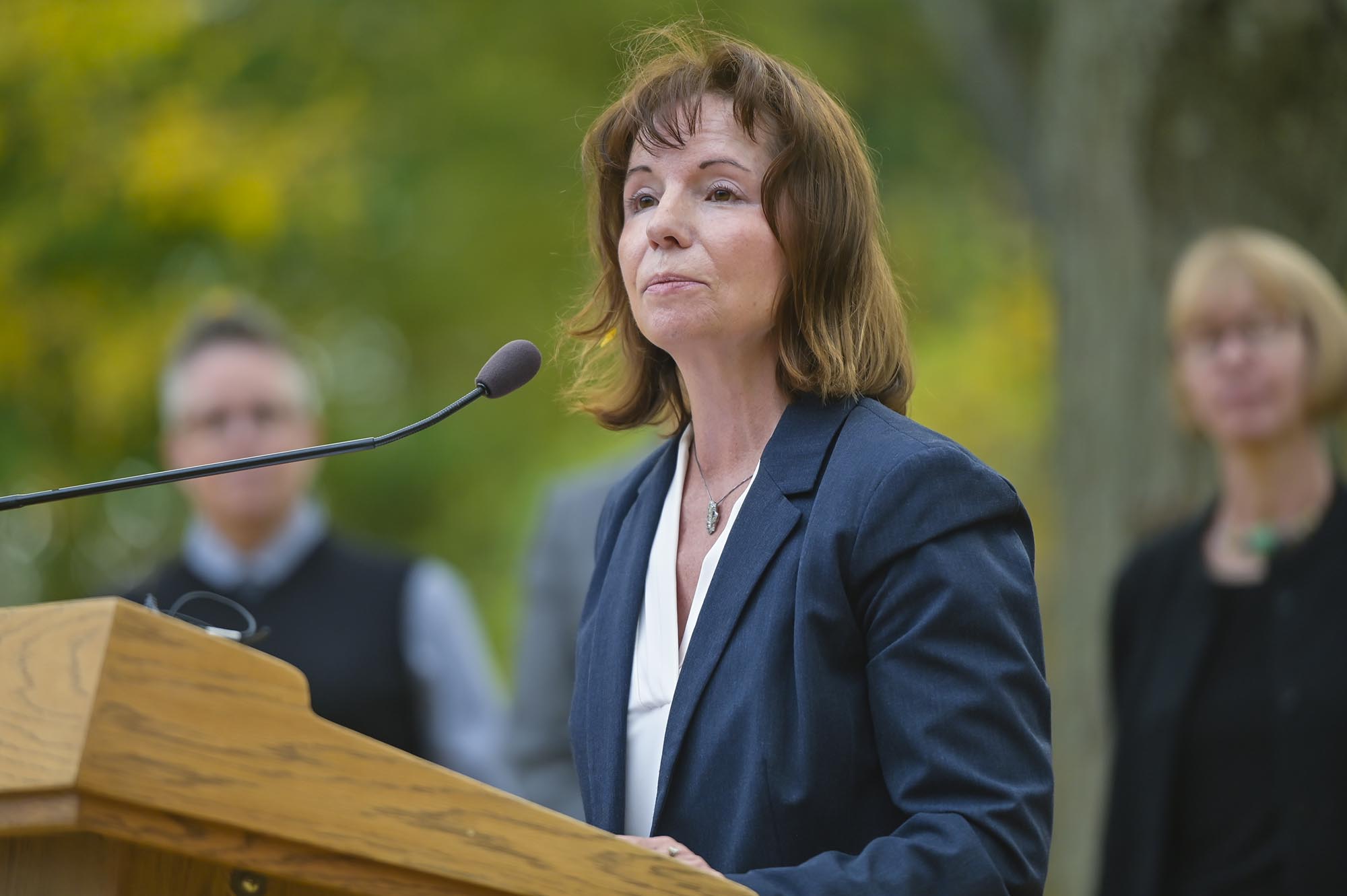 A woman in a blazer speaks at an outdoor podium, with a microphone in front of her and three blurred people in the background.