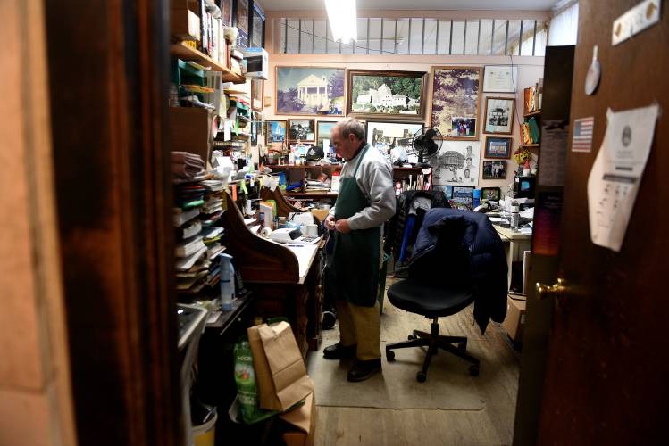 A person in an apron stands in a cluttered office, surrounded by shelves, papers, and framed pictures on the walls.