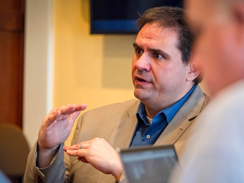 Man in a gray suit gesturing while speaking to another person.