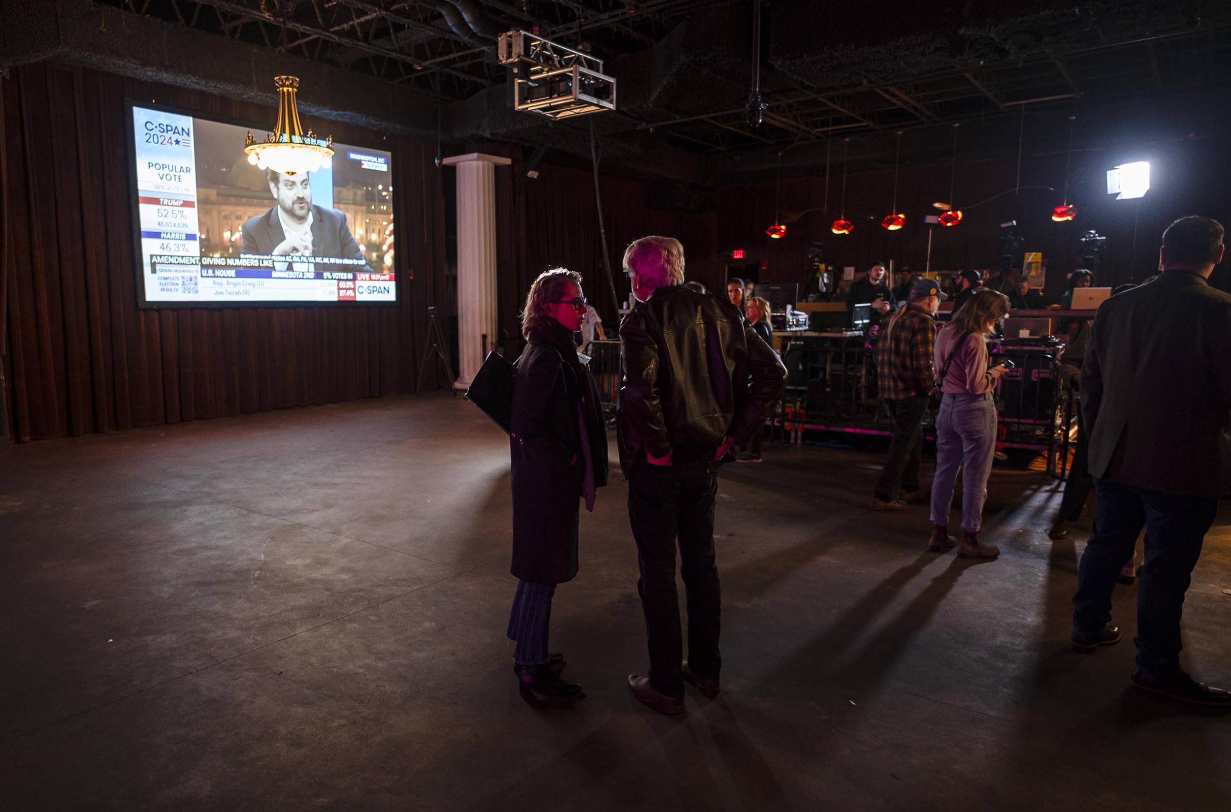 People gather in a dimly lit venue with a large screen displaying news. Some individuals engage in conversation.