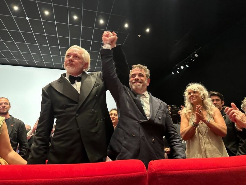 Two men in formal attire stand in a theater, one raising the other's arm in celebration, while people around them applaud.