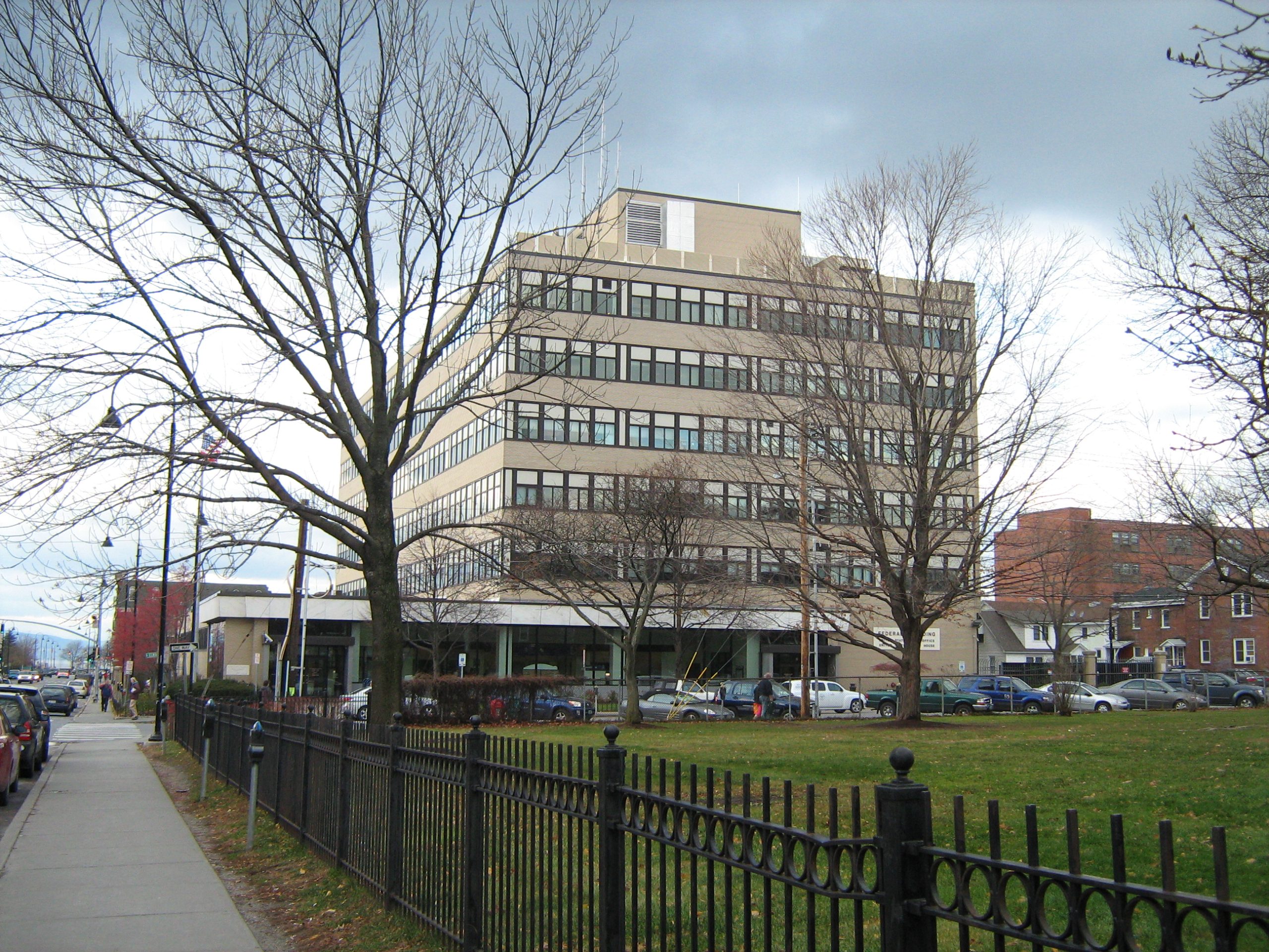A multi-story building with numerous windows is situated along a sidewalk with adjacent trees and a fenced green space in front. Several vehicles are parked nearby. The sky is cloudy.