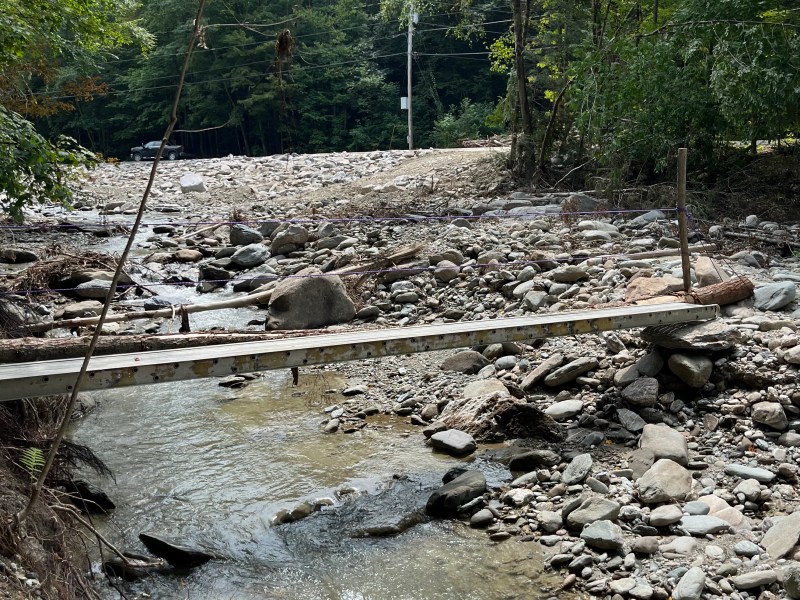A narrow wooden bridge crosses a rocky stream surrounded by trees and shrubbery. The streambed is filled with large stones and debris.