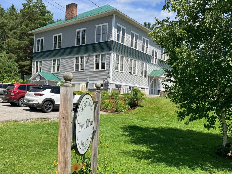 A two-story building with white siding and green trim, identified as "Greensboro Town Offices" by a sign in the foreground. Several cars are parked to the left on a sunny day.