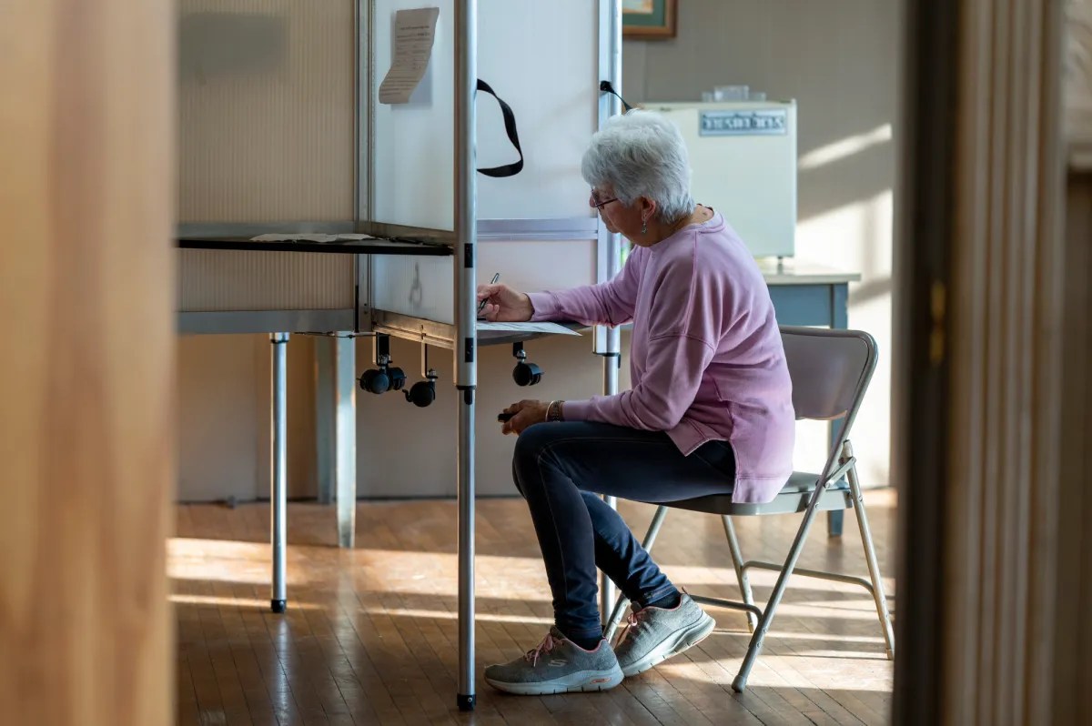 An elderly woman sits in a voting booth, filling out a ballot with a pen.