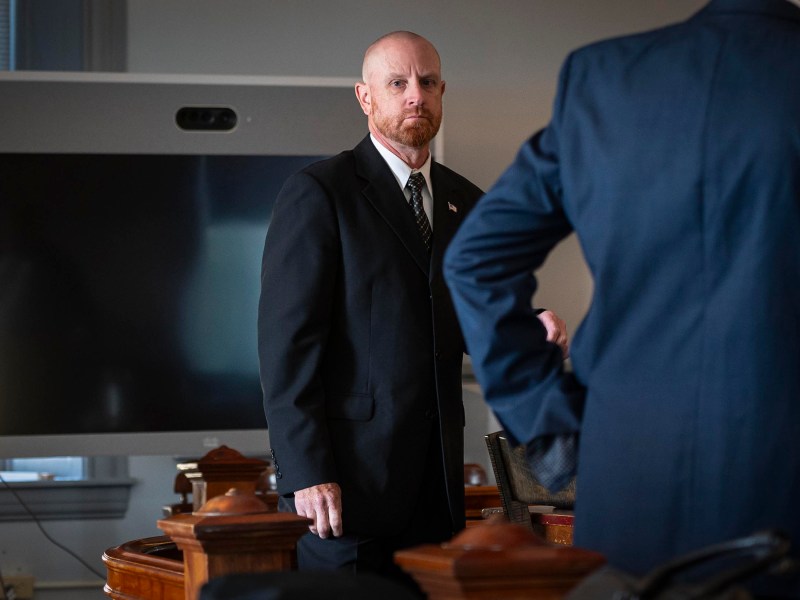 A man in a suit stands in a room with a large screen behind him. Another person, partially visible, also in a suit, stands nearby. The setting appears to be formal or professional.