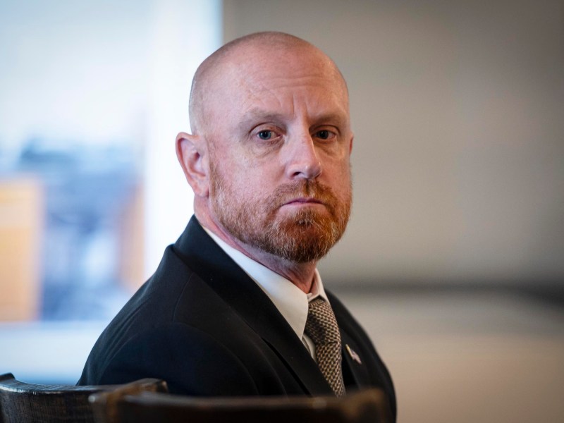 A man with a beard wearing a suit sits in a chair, looking ahead with a neutral expression.