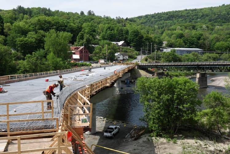 Workers stand on a partially constructed bridge over a river, with another bridge and green, forested hills visible in the background.