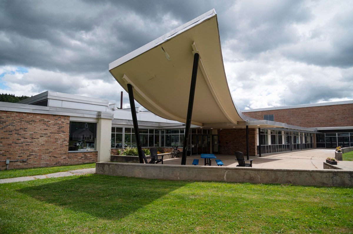 A modern building with a distinctive curved canopy supported by black pillars, brick and glass walls, and a patio area with outdoor seating on a partly cloudy day.