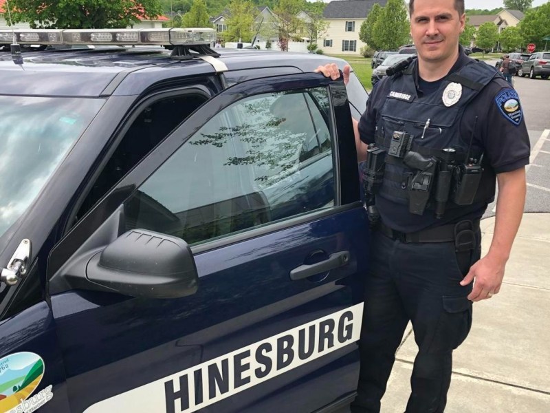 A police officer stands next to a Hinesburg police car parked on a residential street.