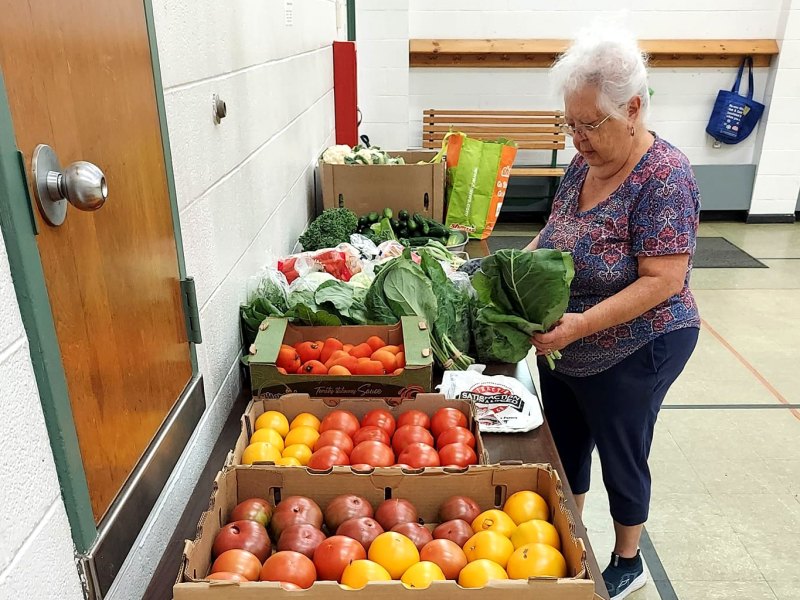 A woman selects leafy greens from a table displaying various fresh vegetables, including tomatoes, in a community center setting.