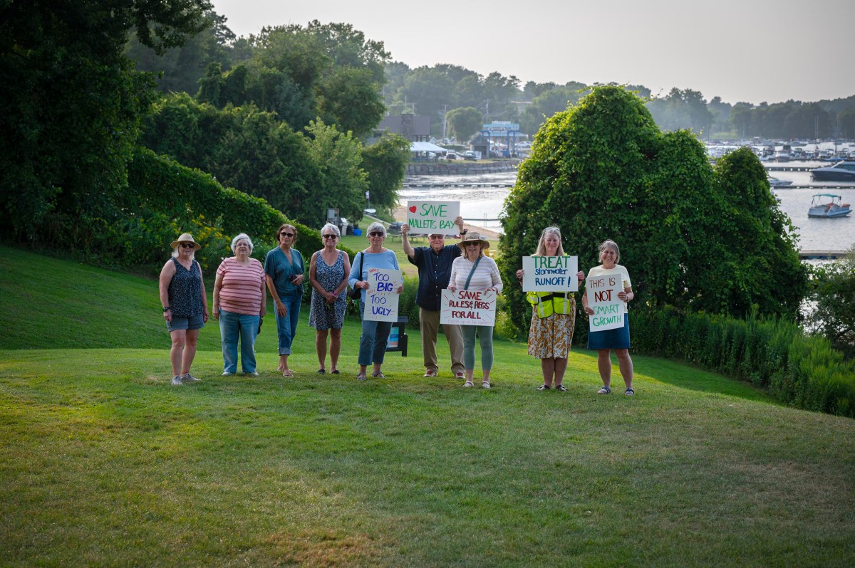 A group of people stand on a grassy slope holding signs with messages about saving Walnut Beach, with trees and a marina in the background.