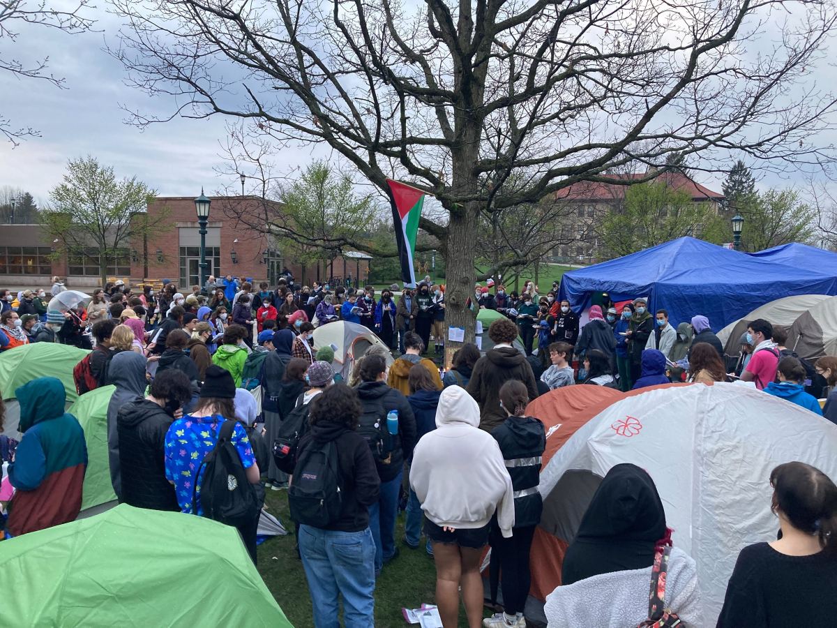 A crowd of people gather around tents and a tree