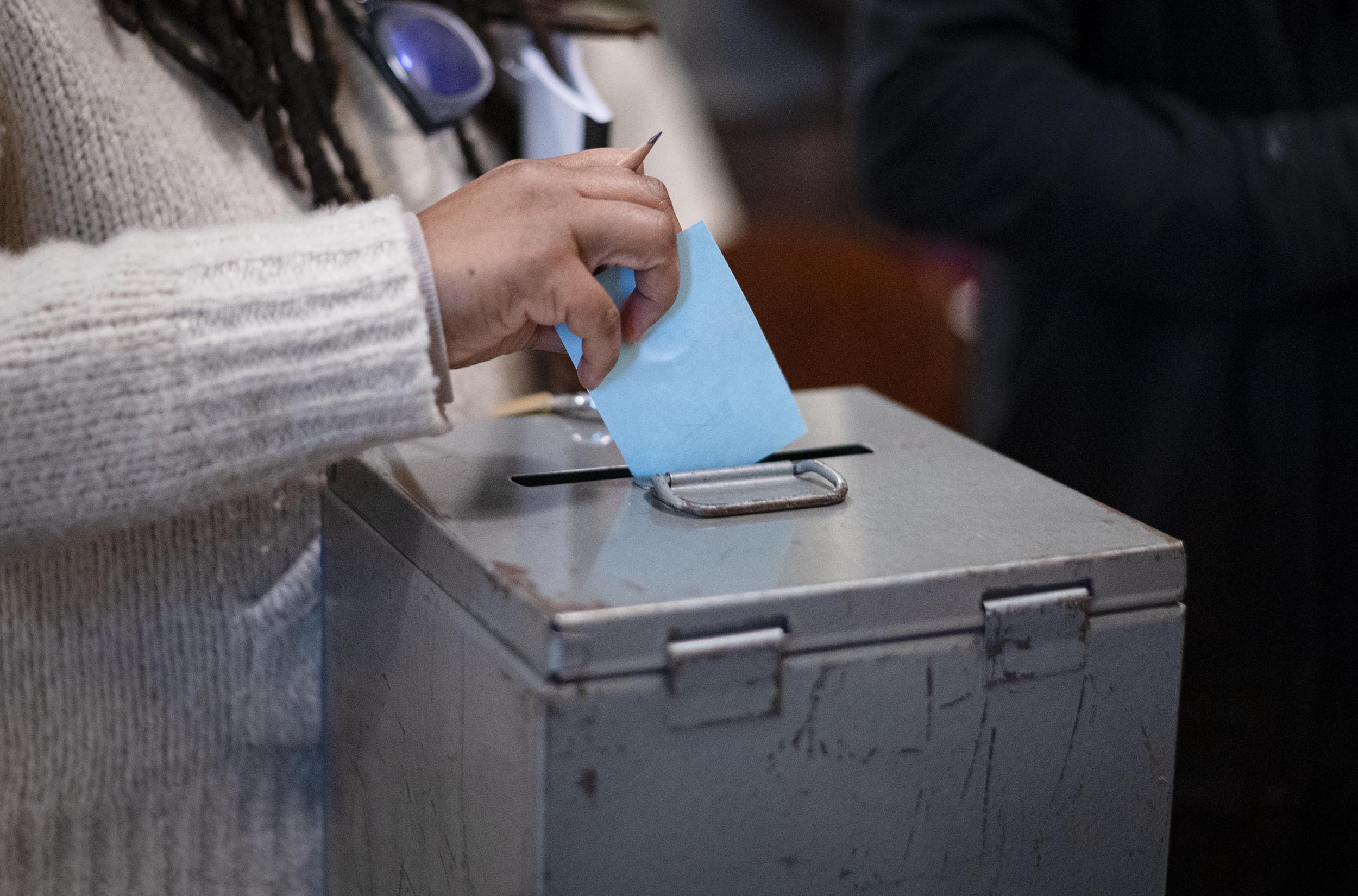 Person placing a blue paper into a gray metal ballot box.