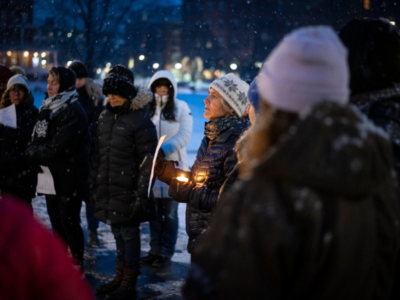 A group of people in winter clothing participate in a nighttime candlelight vigil in a snowy outdoor setting.