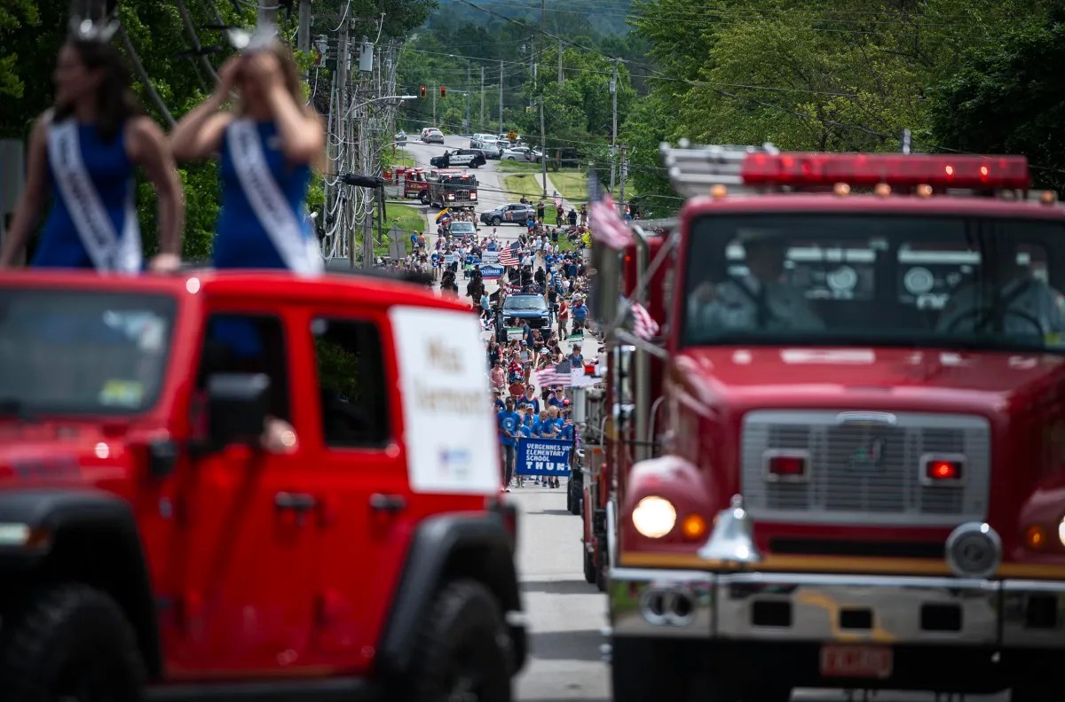 A parade with fire trucks, people walking, and vehicles with passengers in blue sashes takes place on a tree-lined street on a sunny day.