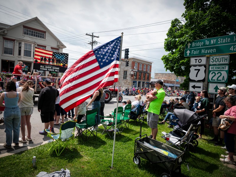 A crowd watches a parade on a street lined with American flags and signposts indicating directions to state routes. A large American flag waves prominently in the foreground.