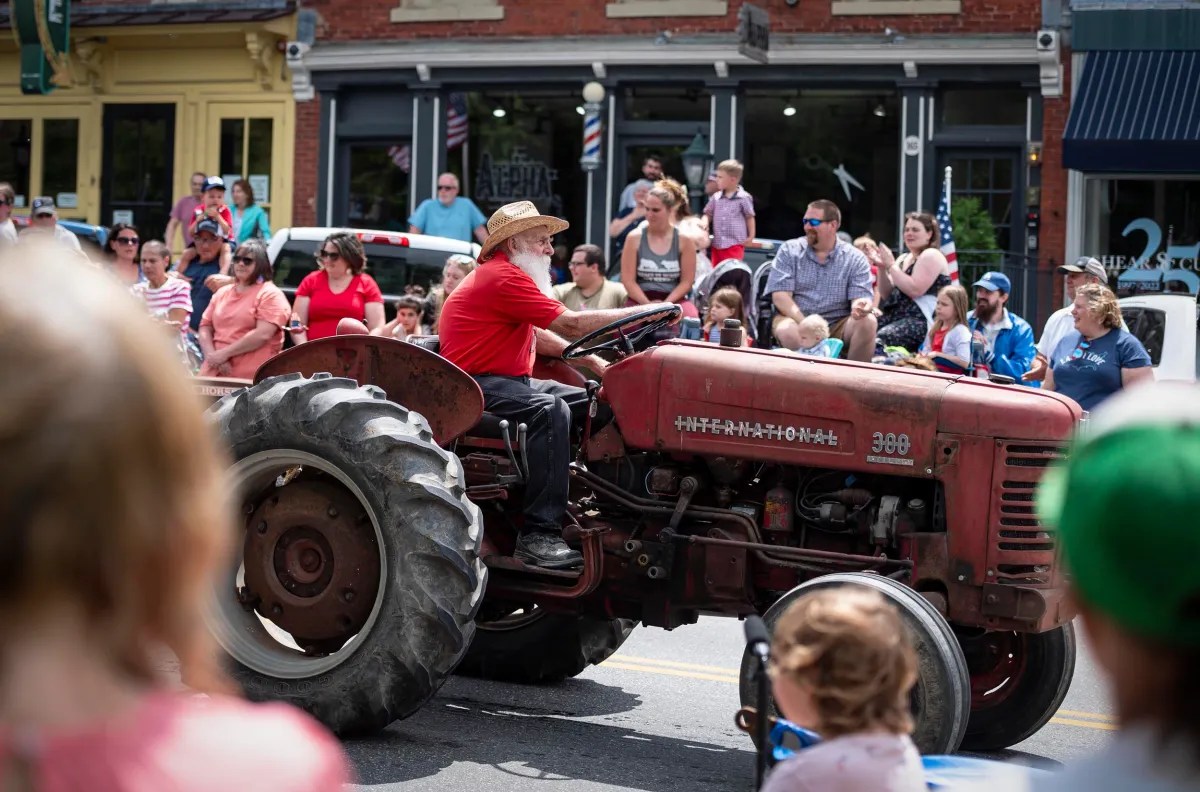 A man in a red shirt and straw hat drives a red tractor down a street during a parade, with a crowd of onlookers on either side.