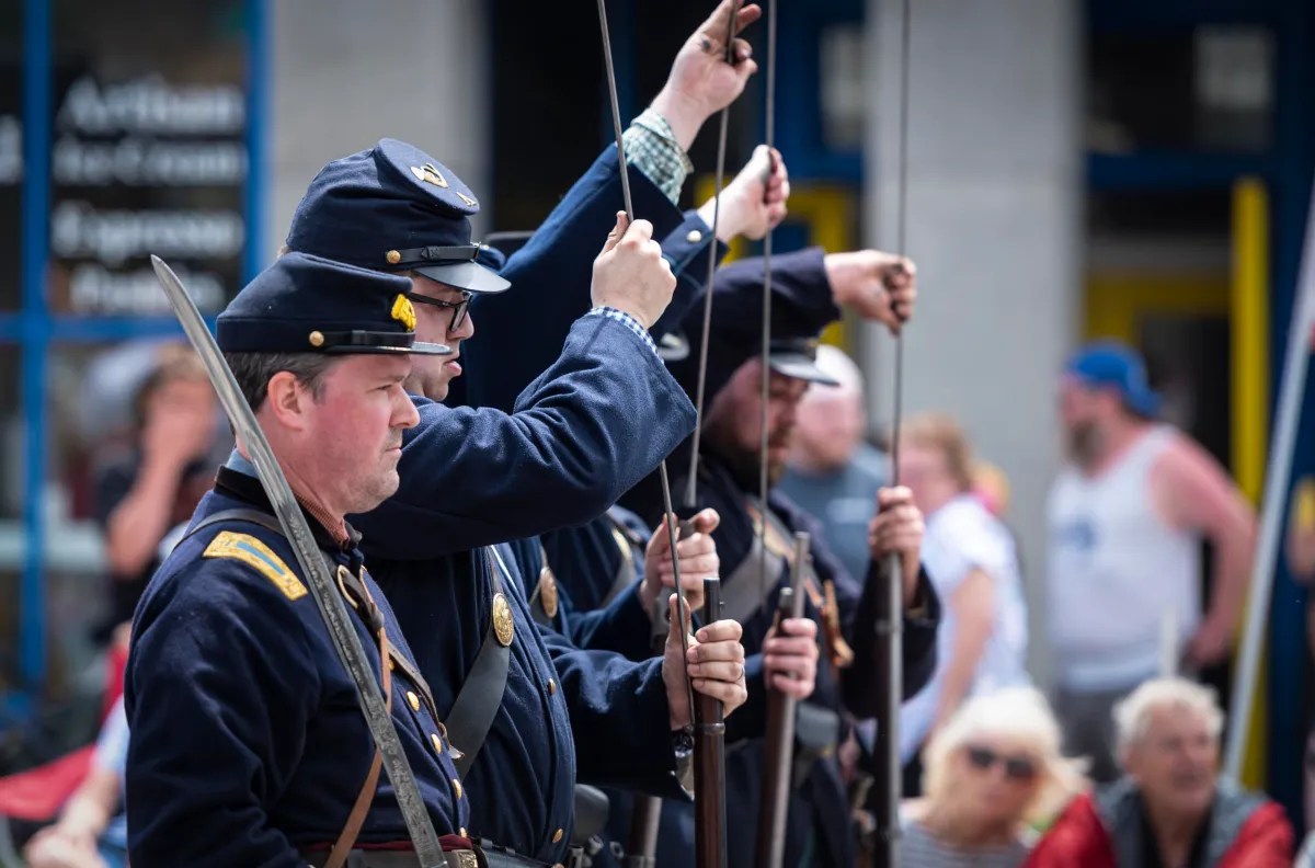 A group of men dressed in blue uniforms and caps hold rifles upright, likely reenacting a historical military scene, with a crowd of spectators in the background.