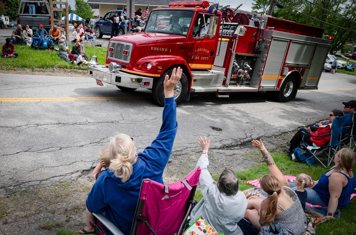 A red fire truck labeled "Engine 1" drives by on a road during a parade as seated people wave at it and watch from the roadside.