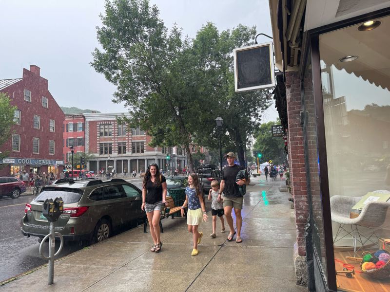 A family walks on a wet sidewalk in a small downtown area on a rainy day, with cars parked along the street and people in the background.
