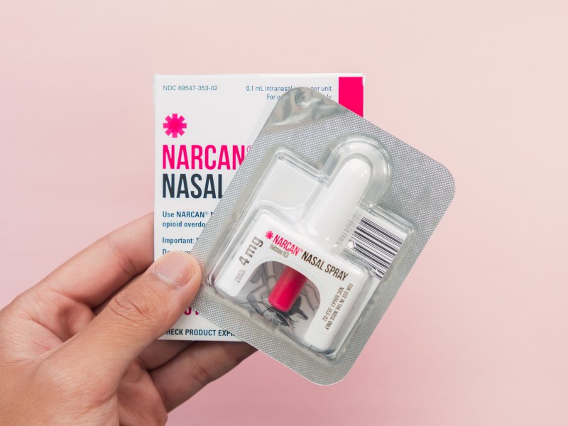 A hand holding Narcan Naloxone against a pink background.