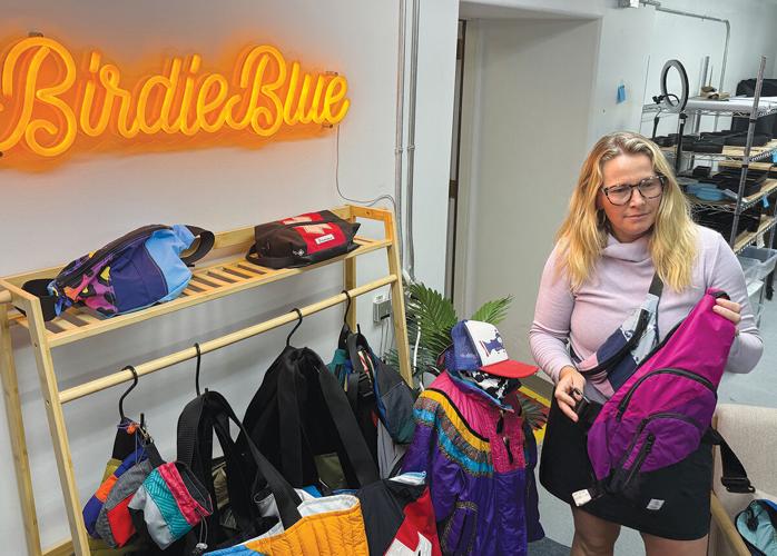 A woman stands in a store next to a rack of colorful bags beneath a neon "BirdieBlue" sign, holding a purple bag and looking at the display.