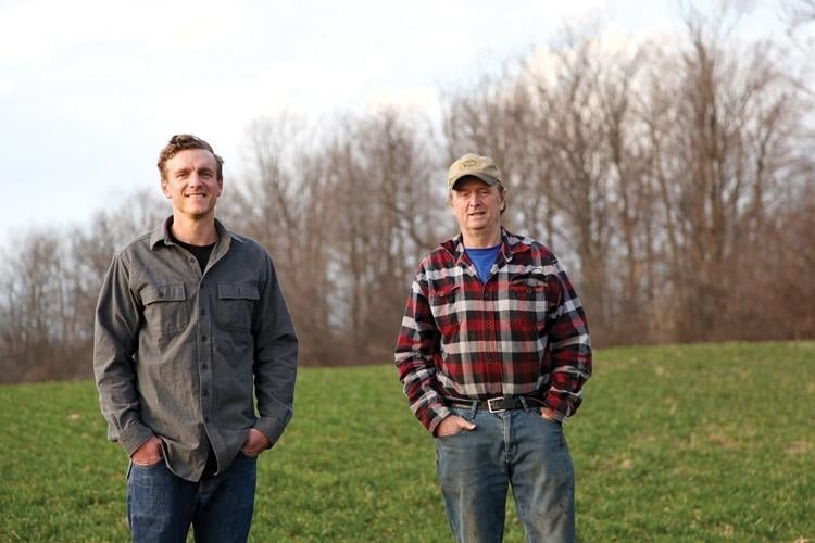 Two men stand in a grassy field with bare trees in the background. They are wearing casual clothes, and one has a cap.