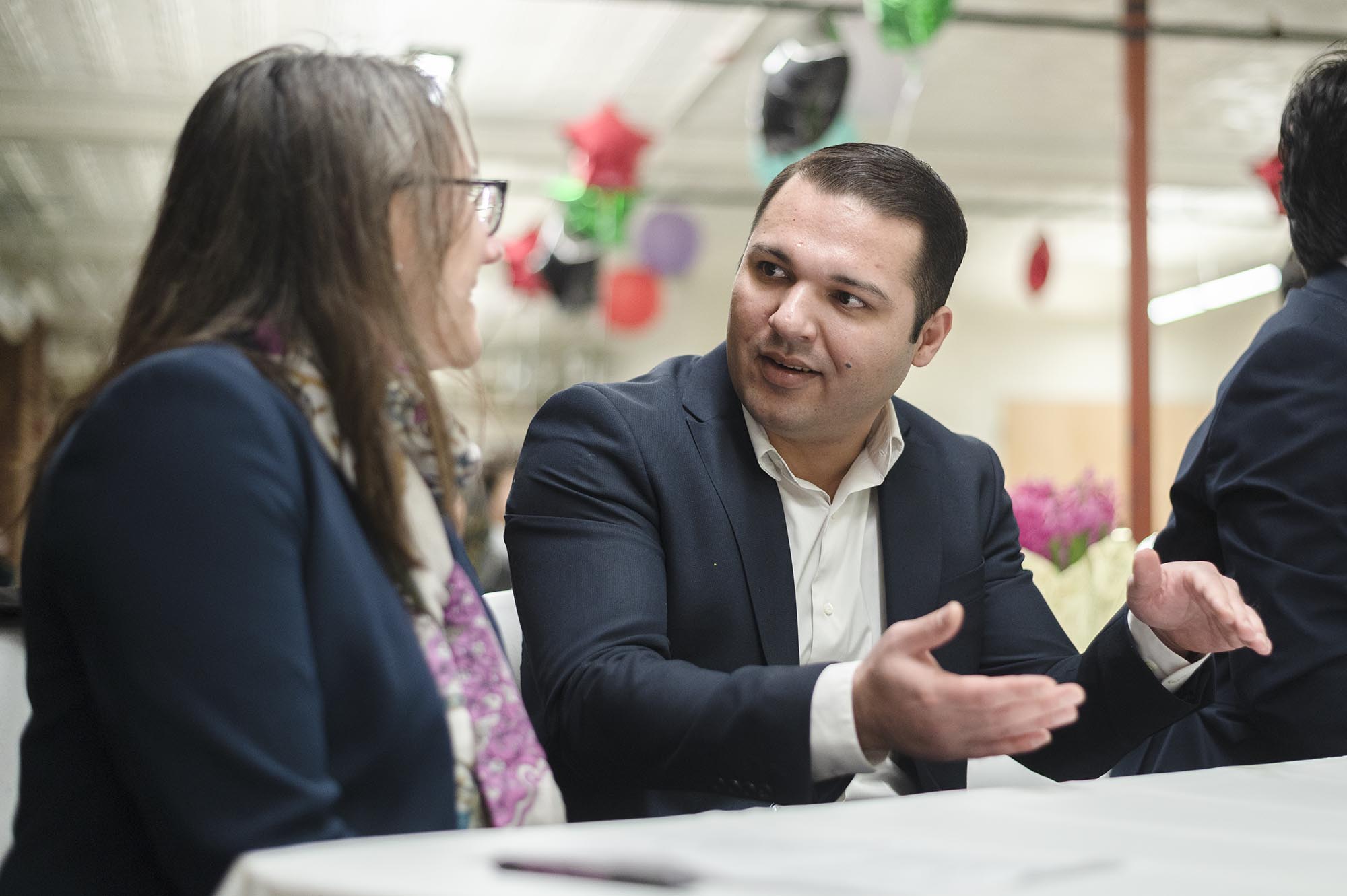 A man in a suit is talking to a woman in a suit who is wearing glasses. They are seated at a table with a white tablecloth. The background is decorated with colorful hanging ornaments.
