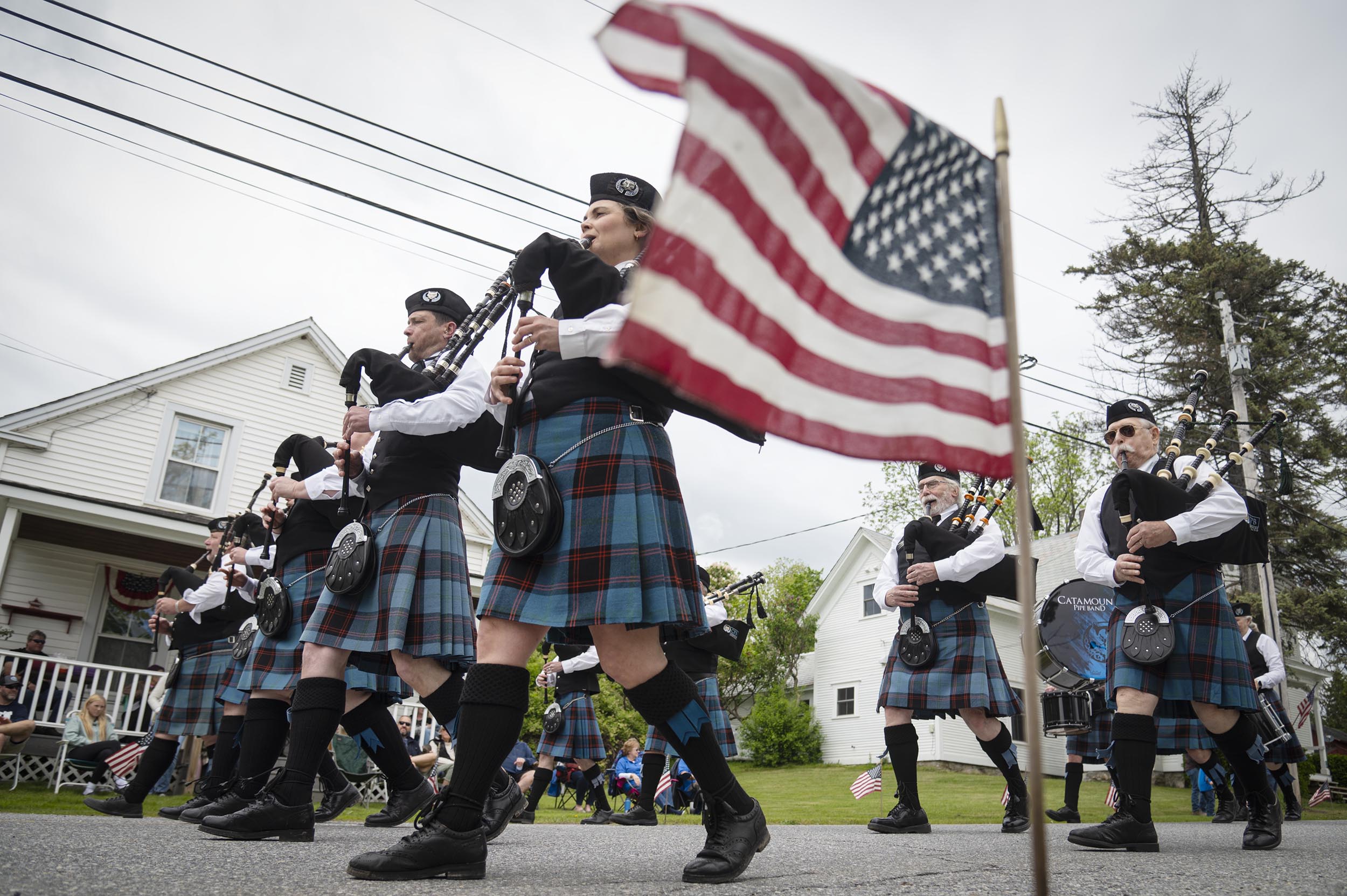 People in matching plaid uniforms play bagpipes while marching in a parade; an American flag is visible in the foreground.