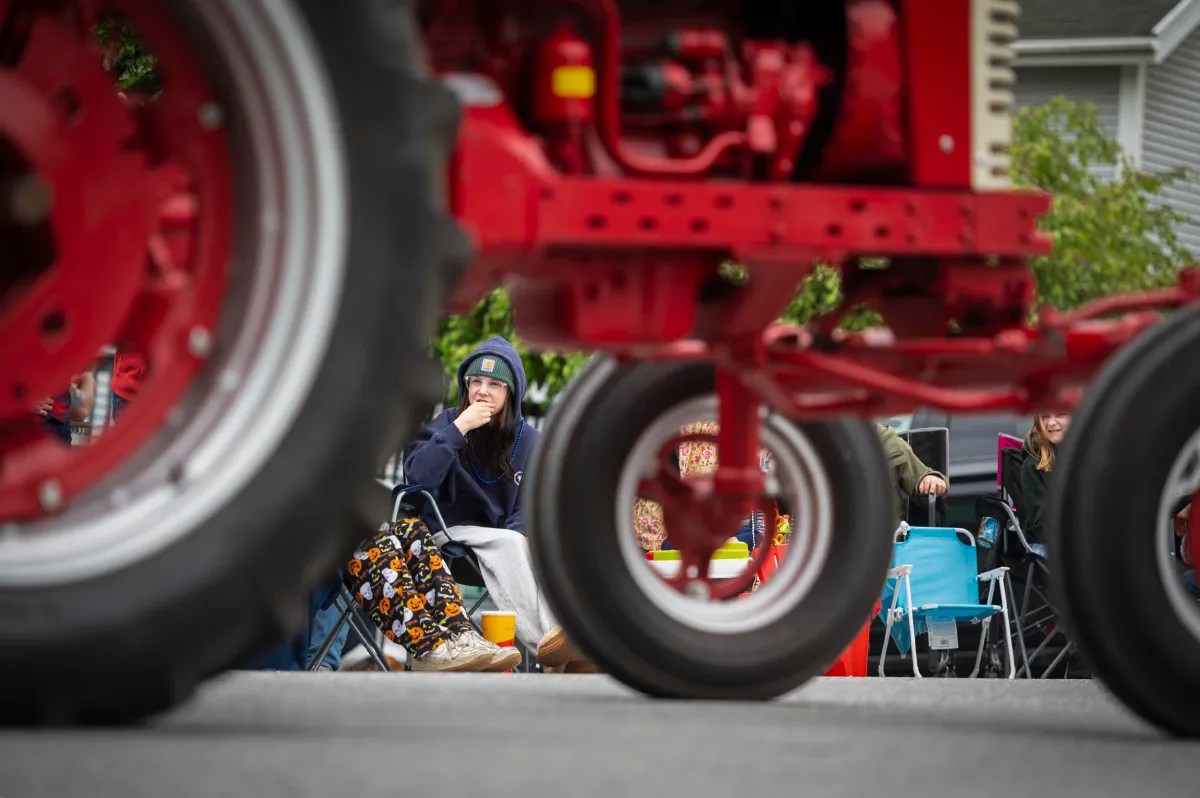 A person sits in a lawn chair watching a parade, partially obscured by the wheels and frame of a red tractor passing by on the street.